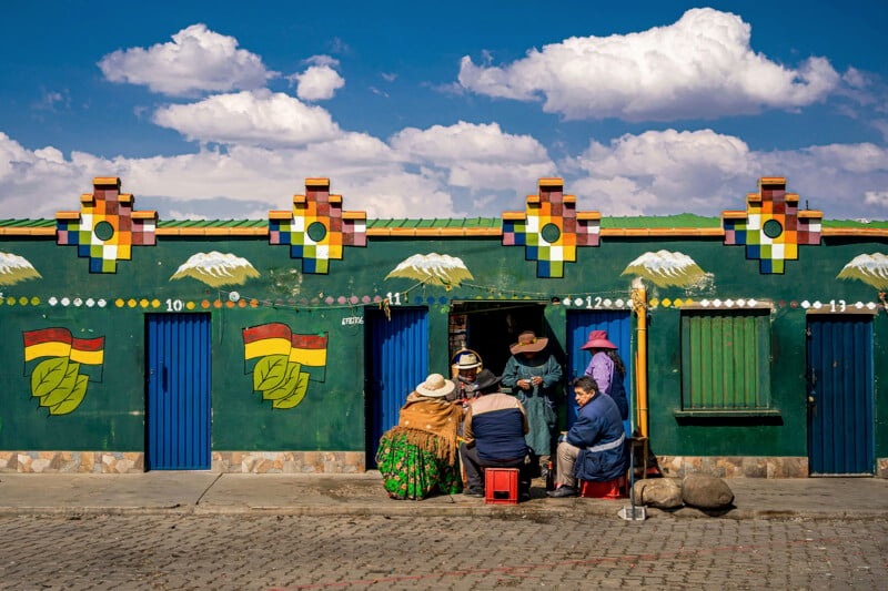 A group of people in traditional clothing sit outside a vibrant green building with colorful geometric and mountain designs. The building features blue doors with painted flags. Fluffy clouds fill the blue sky above.