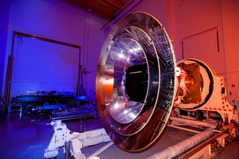 A large, reflective dish antenna is set up in a dimly lit room with blue and pink lighting. The antenna is mounted on a white mechanical base, and wires and equipment are visible in the background, suggesting a high-tech facility.