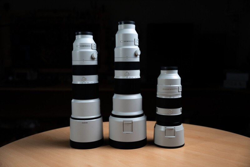 Three white camera lenses with black accents are displayed upright on a wooden table against a dark background. The lenses vary in size, with the tallest on the left and the shortest on the right.