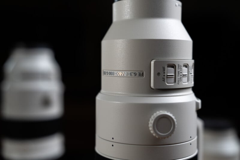 Close-up of a white camera lens with switches for focus settings. The text "4.5-5.6/100–400 GM OSS" is visible. Another blurry lens is in the background against a dark backdrop.