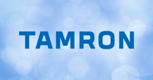 The image features the word "TAMRON" in bold blue letters against a light blue background with soft, circular bokeh effects.