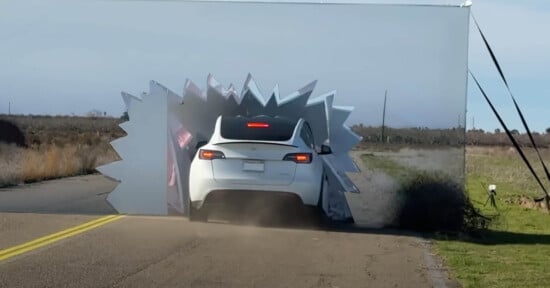 A white car is crashing through a screen that imitates a zigzag pattern or a starburst. The screen has partially collapsed around the car. Dust is visible around the vehicle as it drives down a rural road.