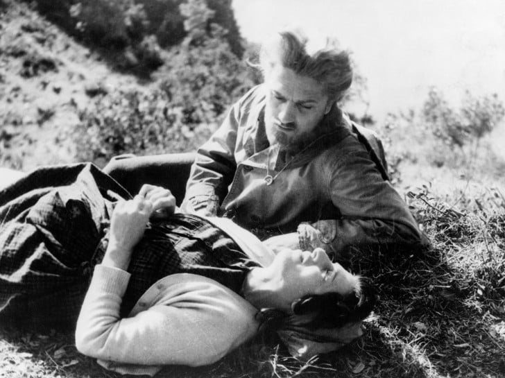 A man with a beard leans over a woman lying on the grass, outdoors. They appear engaged in a serious conversation. The background is blurred, suggesting they are in a natural setting. Both wear mid-20th-century clothing.