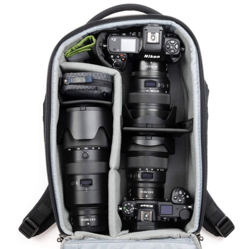 Open camera backpack showing organized lenses and camera bodies. Labeled Nikon lenses and cameras are neatly arranged, with compartments for accessories. The interior is padded and designed for secure transport.