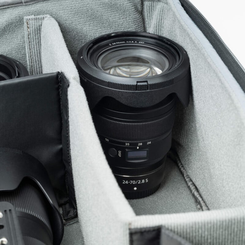 A zoom lens labeled "24-70/2.8 S" is neatly placed in a gray, padded camera bag compartment. Another lens is partially visible next to it in the bag. The focus is on the lens’s details and the structured interior of the bag.