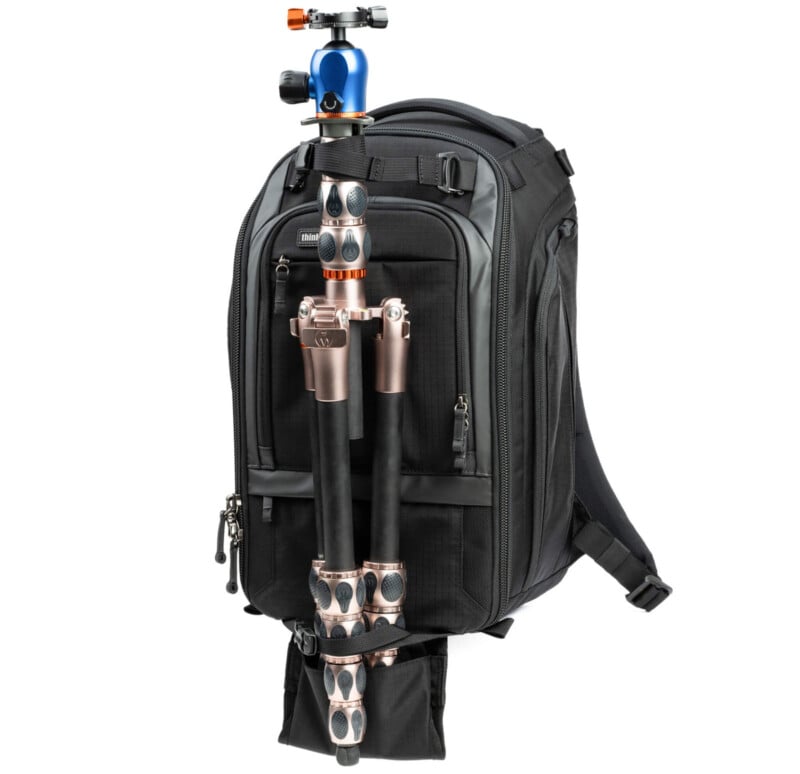 A black backpack with multiple compartments has a tripod attached to its front using straps. The tripod features black legs with silver and copper accents and a blue-and-orange ball head at the top.