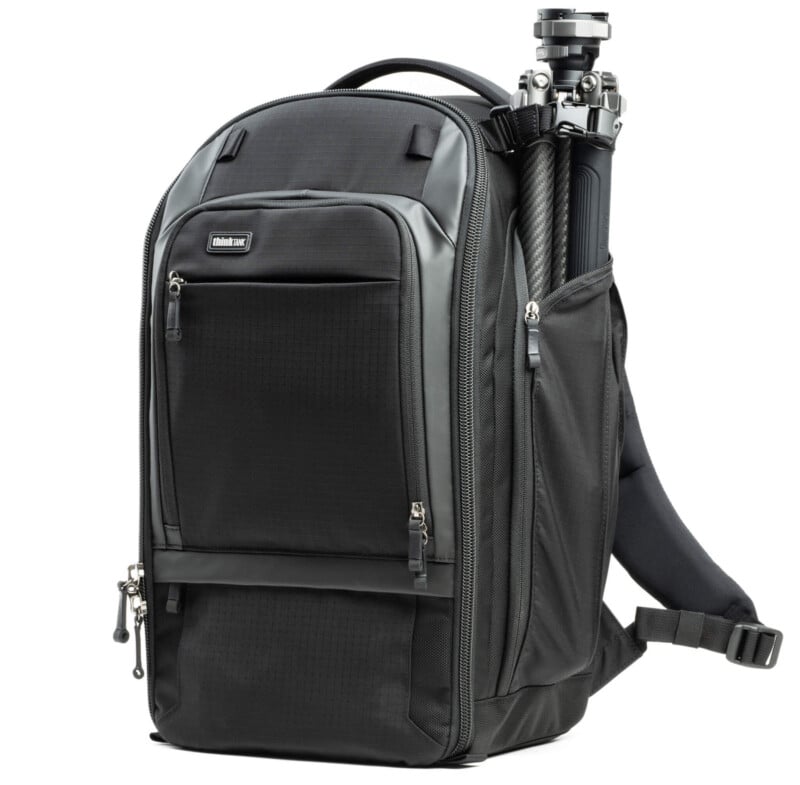 Black camera backpack with multiple zippered compartments and a tripod attached to the side. The backpack features adjustable padded straps and a top handle, designed for carrying photography equipment.