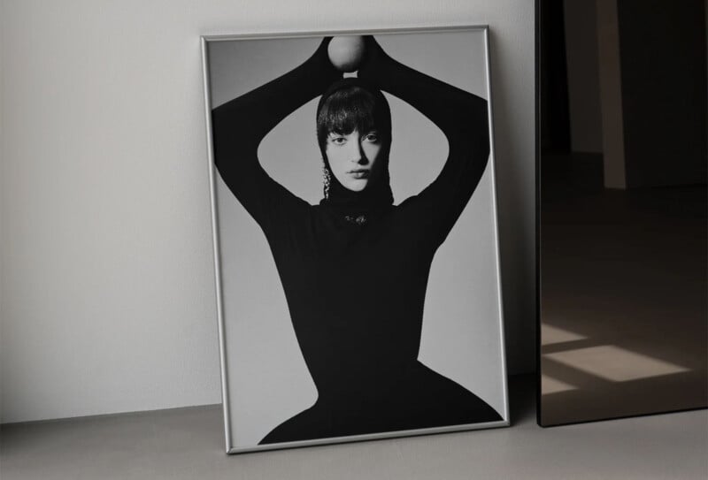 A framed black and white photograph of a person in a fitted black outfit, posing with hands above their head, leaning against a light-colored wall. The setting is minimal, with a mirror reflecting part of the room.