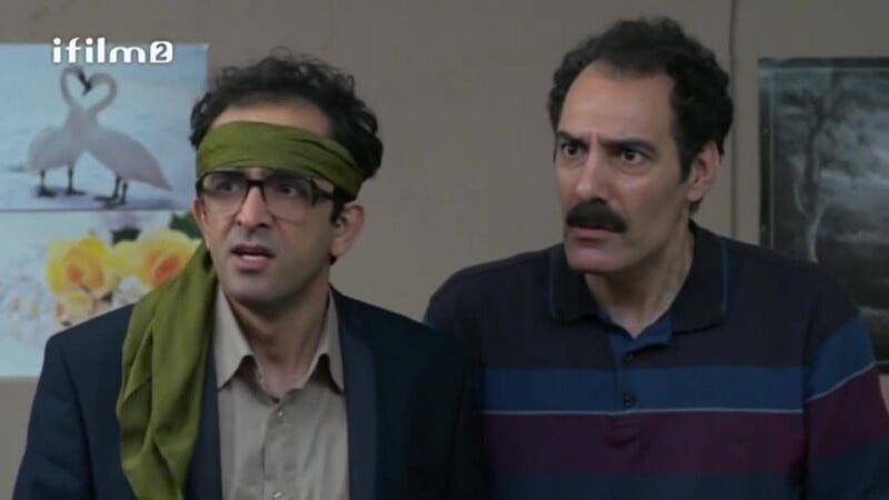 Two men look concerned indoors; one wears glasses, a suit, and a green headband, while the other has a mustache and is dressed in a striped sweater. Swans and flower pictures are on the wall behind them.