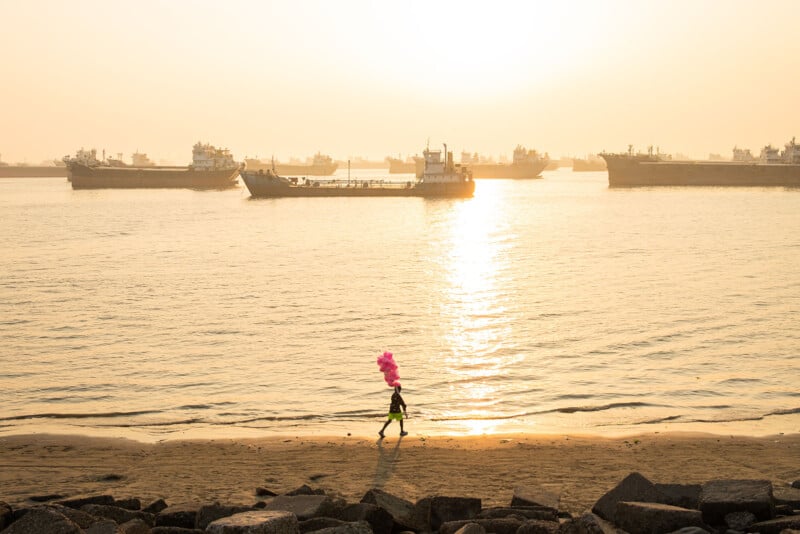 A person walks along a sandy beach at sunset, holding bright pink cotton candy, with large ships anchored in the calm water and sunlight reflecting off the sea. Rocks are visible in the foreground.
