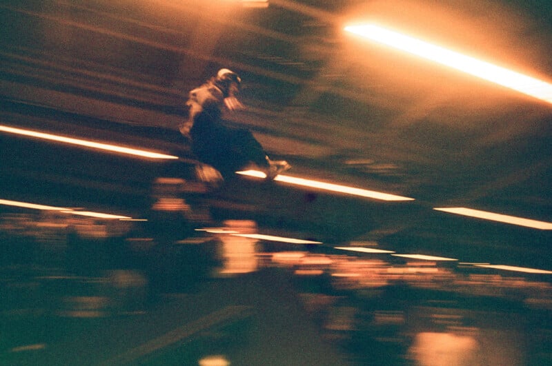 Blurred image of a skateboarder mid-jump in an indoor skate park, with bright lights streaking across the scene, capturing a sense of motion and energy. The background is filled with dimly lit silhouettes of bystanders.