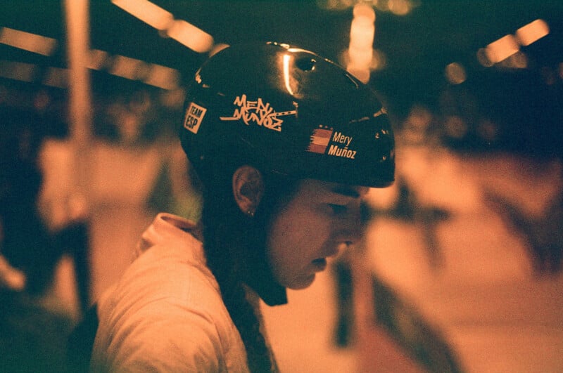 A person wearing a helmet with stickers is in focus, surrounded by a blurred, dimly lit indoor environment. The atmosphere is warm with a mix of orange and green hues, creating a nostalgic effect.