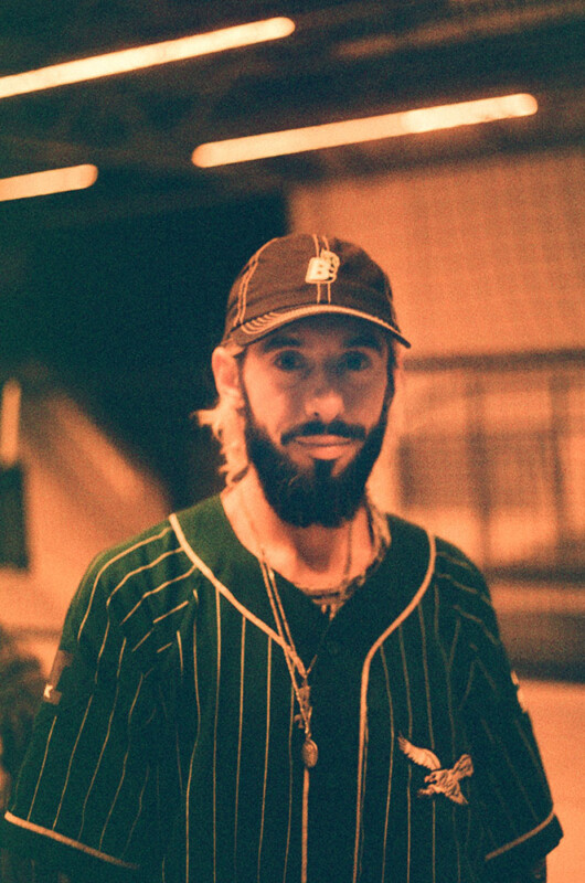 A person with long hair and a beard wearing a baseball cap and a striped baseball jersey. They have several necklaces. The image has a warm, vintage filter with blurred background featuring lights and a structure.