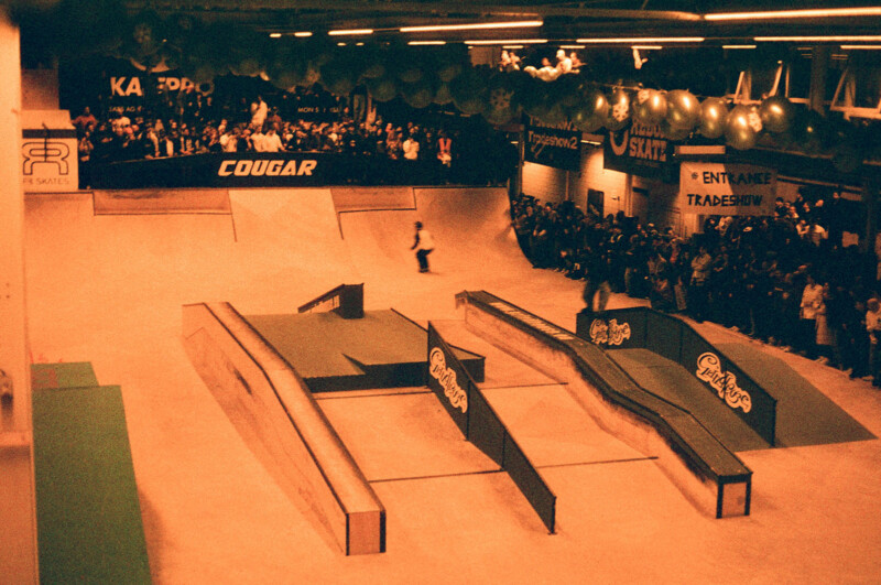 A vintage-style photo of an indoor skatepark competition. A skater performs on ramps and rails while a large crowd watches. The venue is decorated with banners and balloons, and the image has a reddish-brown tone.