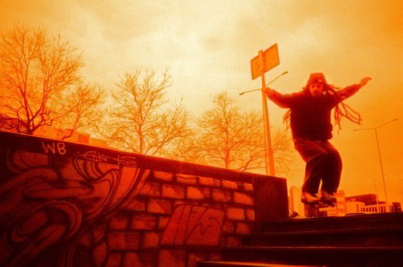 Person with dreadlocks skateboards down stairs under an orange sky. Bare trees and graffiti on a brick wall in the background create a dynamic urban scene.