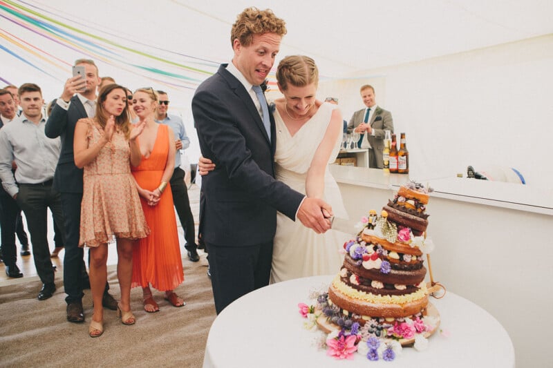 A bride and groom, smiling, cut a tall, colorful wedding cake together as guests watch and take photos in a bright, decorated venue with rainbow streamers.