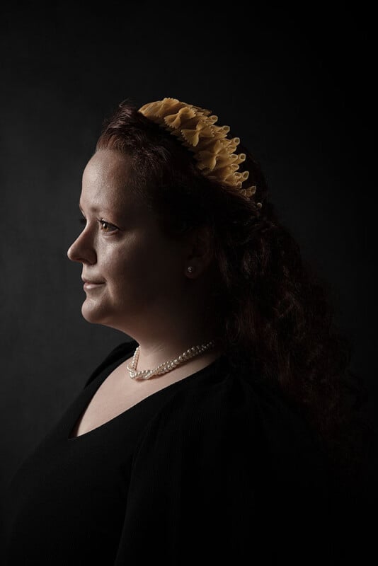 A woman with curly brown hair wears a yellow headband and pearl necklace, dressed in black, facing left in profile against a dark, softly lit background.