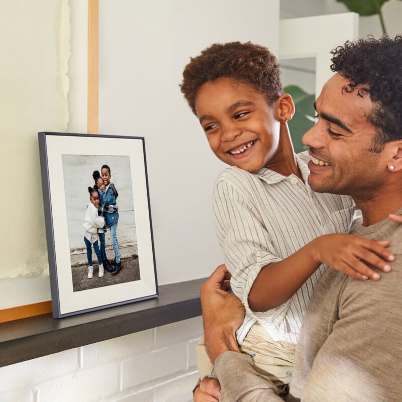 A smiling man holds a happy young boy near a mantelpiece, where a framed photo of two children—one hugging the other—is displayed. The setting appears warm and homey with simple decor.