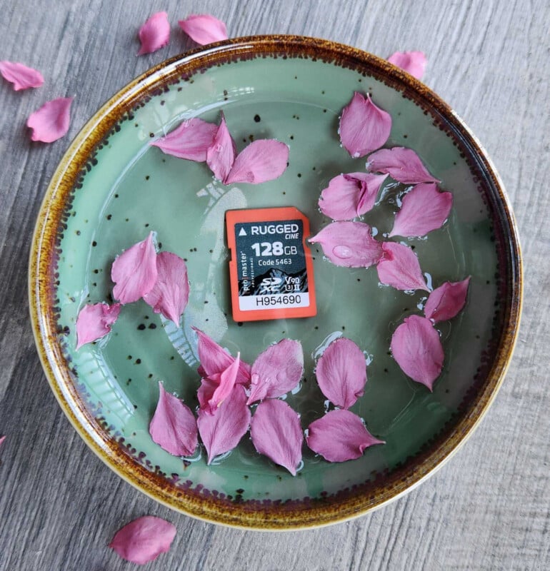 A ceramic bowl with green glaze and brown rim sits on a wooden surface, filled with water, pink flower petals, and a 128GB memory card floating on top.