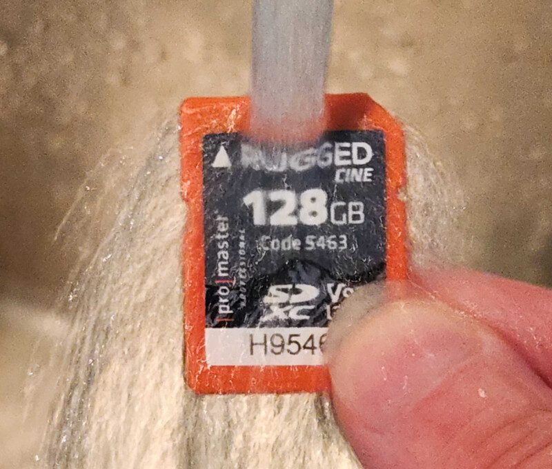 A person holds a 128GB SD card labeled "RUGGED Line" under running water, demonstrating its water resistance.