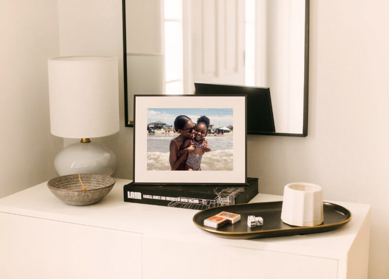 A white dresser topped with a lamp, a bowl, a tray with a candle and matches, and a framed photo of two people smiling at the beach, with a mirror and white walls in the background.