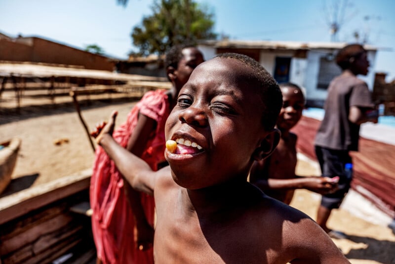 A smiling boy stands outdoors in the sunlight, playfully sticking out his tongue with a small round object on it. Other children and simple buildings appear in the blurred background.