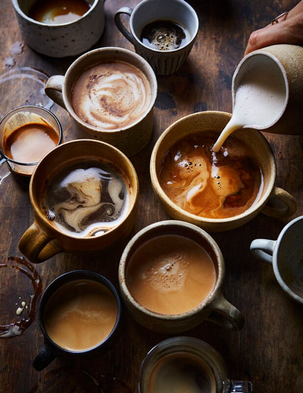A variety of ceramic mugs filled with different types of coffee are arranged on a wooden table. Milk is being poured into one mug, creating swirling patterns. Some cups have spilled coffee around them.