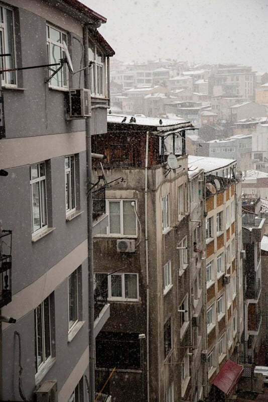 Snow falls on a dense cluster of multi-story apartment buildings in a city, with overcast skies and rooftops dusted with snow. The scene appears cold and quiet, showing windows and balconies along the buildings.