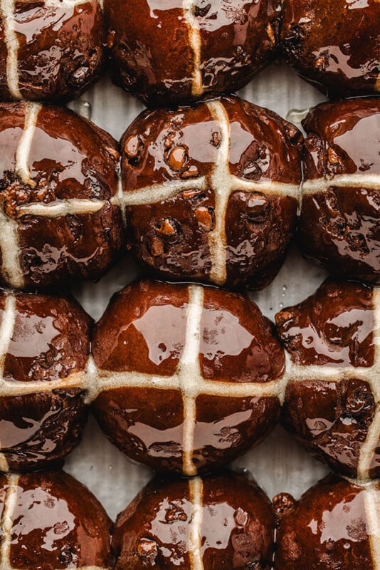 Close-up of shiny, freshly baked chocolate hot cross buns arranged in rows. Each bun has a white cross on top, and the buns have a glossy, glazed surface.