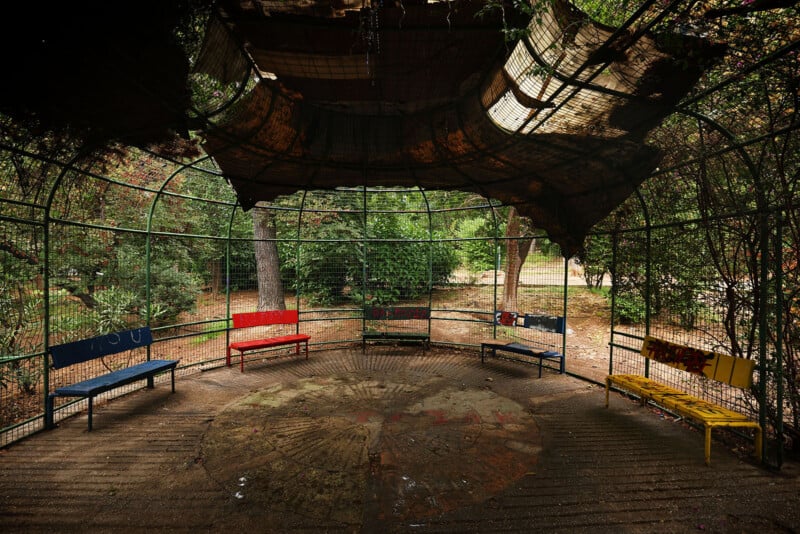 A round, fenced outdoor structure with a partially covered roof and several colorful benches arranged in a circle. Trees and greenery surround the space, and sunlight filters through the canopy above.