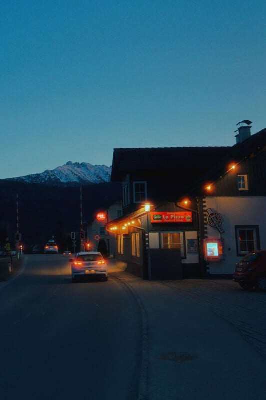 A car drives past a brightly lit pizza restaurant at dusk. The sky is clear, and snow-capped mountains are visible in the background. The street is calm with minimal traffic.