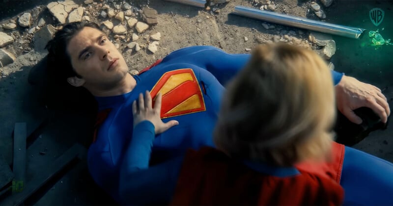 A person in a superhero costume with a large "S" emblem lies on the ground, surrounded by debris. Another person in a red cape is kneeling beside them, placing a hand on their chest. The scene appears dramatic and intense.