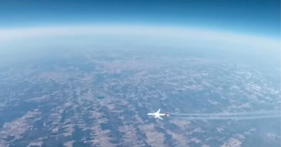 A commercial airplane flies high above a patchwork landscape, leaving a visible contrail behind, with the Earth’s curvature and blue sky in the background.