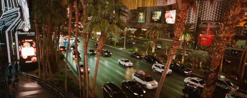 Busy nighttime scene on a city street with tall palm trees and colorful illuminated signs. Several cars drive along the road beside a sidewalk lined with people. Bright, modern buildings glow with vibrant lights and advertisements.