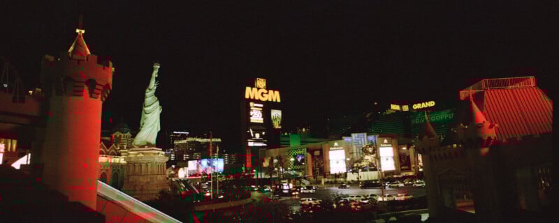 Night view of the Las Vegas Strip featuring the illuminated replica of the Statue of Liberty, MGM Grand building, and various brightly lit hotel and casino signs. Traffic is visible on the street below.