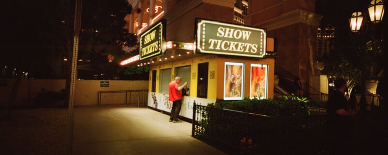 A small kiosk labeled "Show Tickets" is illuminated at night, surrounded by bright signs and posters. A person in a red shirt stands at the kiosk's counter. The scene includes plants and soft street lighting.