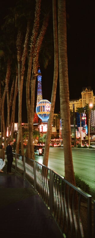 Night scene of a street with palm trees and a brightly lit globe and tower structure in the background. People walk along the sidewalk, and cars drive by on the road. The atmosphere is lively with colorful lights.