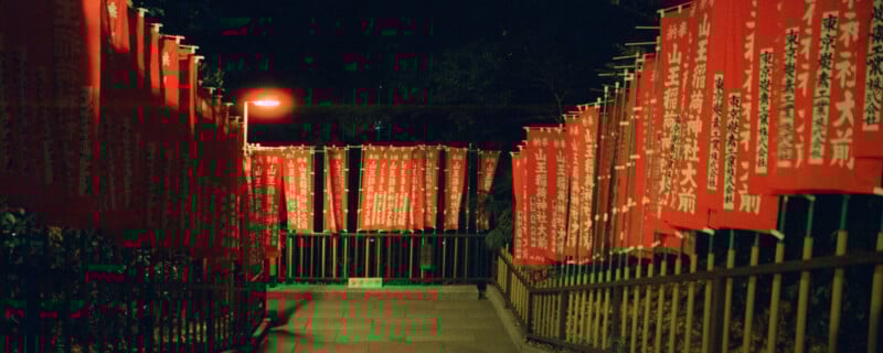 A dimly lit pathway at night is flanked by rows of vibrant red banners with white Japanese script. The banners are attached to poles, creating a tunnel-like effect, with a single streetlamp casting a warm glow.