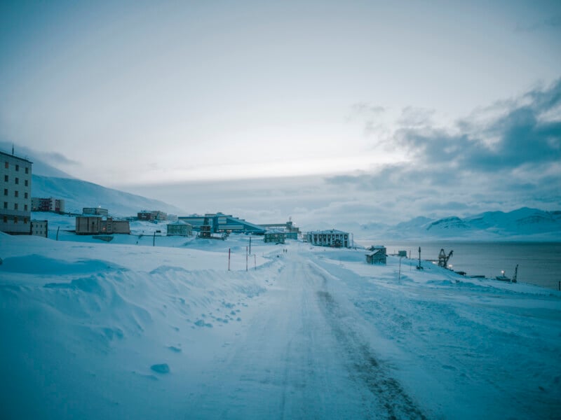 A snow-covered road leads to a small Arctic town with buildings on either side, surrounded by snowy mountains and water under a cloudy, pale sky.