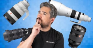 A man with gray hair and beard, wearing a black shirt, stands thoughtfully with his finger on his lips. Four large camera lenses float around him against a blue pixelated background.