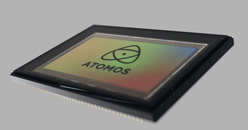 A close-up of a computer chip with a multicolored reflective surface. The black border surrounds the rectangular chip, featuring an atomic symbol and the word "ATOMOS" on its surface, against a plain gray background.