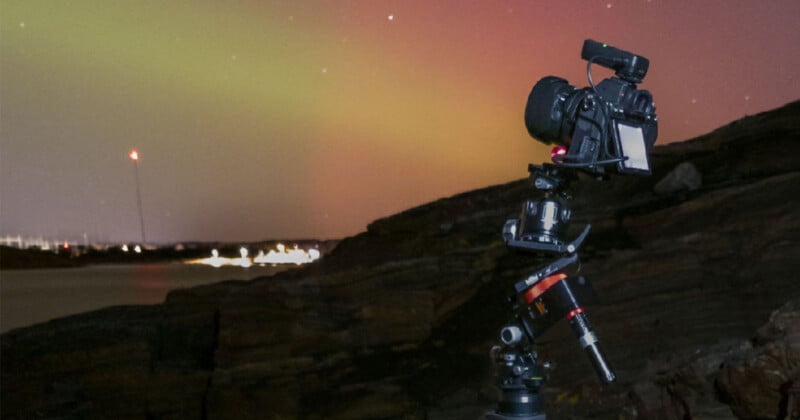 A camera mounted on a tripod is set up on rocky terrain at night, capturing a sky illuminated by green and reddish aurora lights, with distant city lights visible near the horizon.