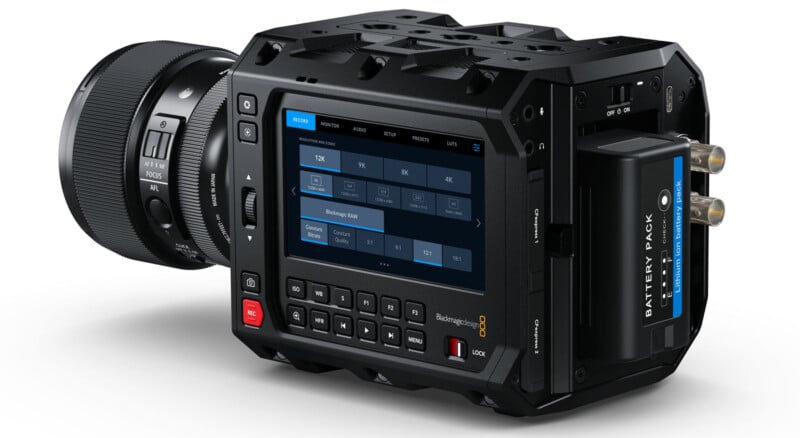 A high-end digital cinema camera with a lens attached, featuring a touchscreen display showing resolution settings. The camera has a rugged design with multiple buttons and a battery pack on the right side.