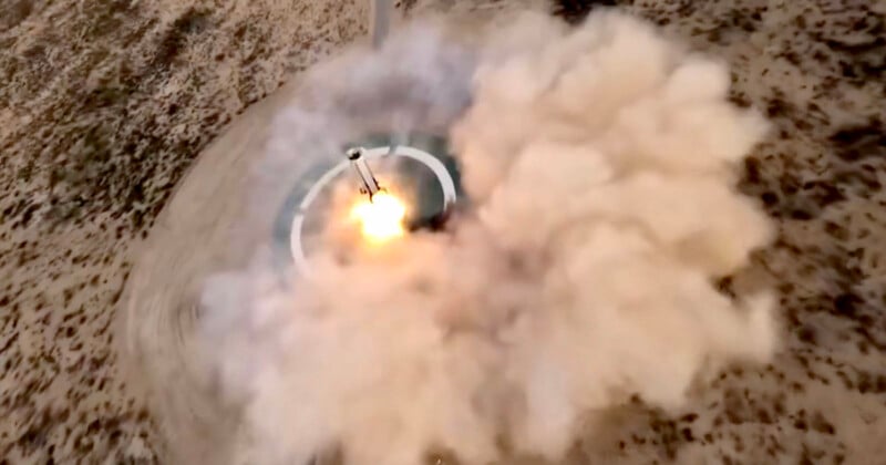A rocket launches from a circular pad in a desert area, producing bright flames and a large cloud of smoke that spreads over the surrounding sandy landscape.