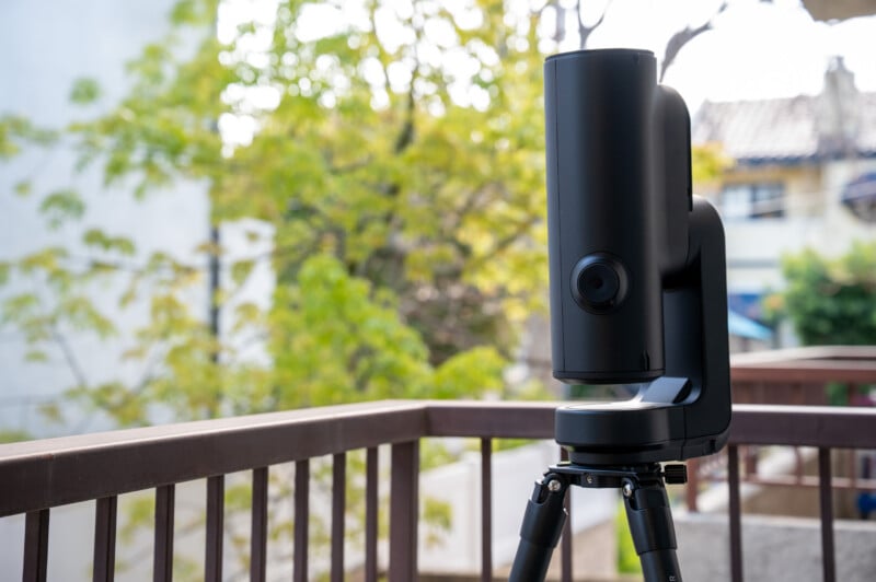 A modern black telescope or camera mounted on a tripod stands on a balcony with trees and buildings blurred in the background.