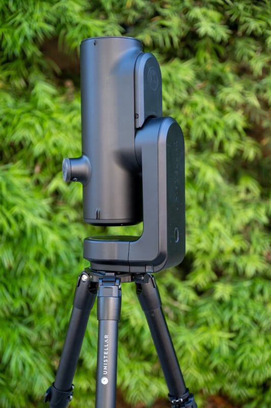 A modern black Unistellar telescope is mounted on a tripod, standing outdoors in front of lush green foliage.