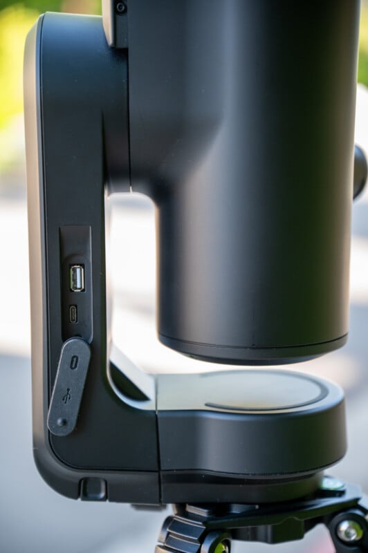 Close-up of a black electronic device mounted on a tripod, showing USB and micro-USB ports and a power button on the side. The background is blurred with natural light.