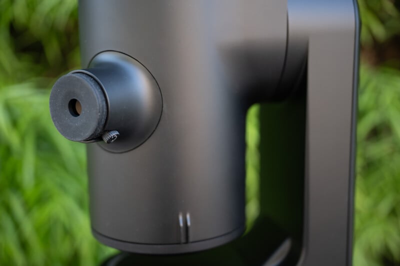 Close-up of a matte black cylindrical device with a protruding round knob and a small screw, set against a blurred background of green leaves.