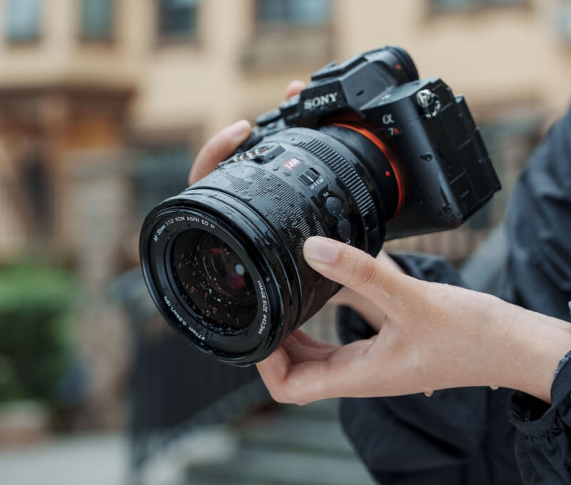 A person holding a Sony Alpha 7R IV camera with a large lens, outdoors. The camera and lens have water droplets on them, suggesting rainy or wet conditions. The background is blurred.