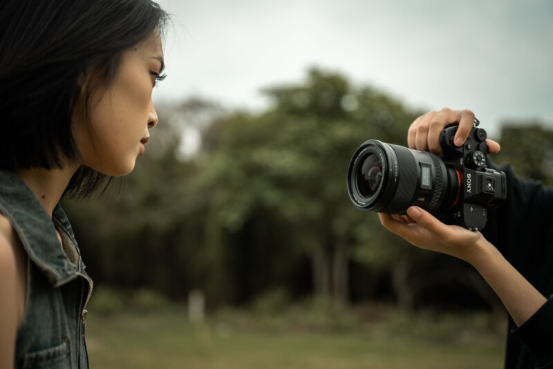 A person holds a Sony camera, aiming it at a woman with short black hair who is looking to the side. The background is blurred greenery, suggesting an outdoor setting.